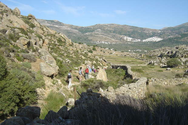 Hikers walk along a narrow trail surrounded by rocky hills and sparse vegetation. In the background, mountains and a distant village are visible under a clear blue sky.